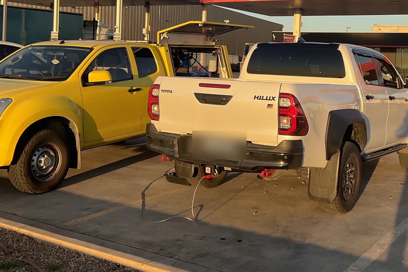 Yellow service ute parked beside a white Toyota Hilux at a servo bowser, hoses laid out on the forecourt