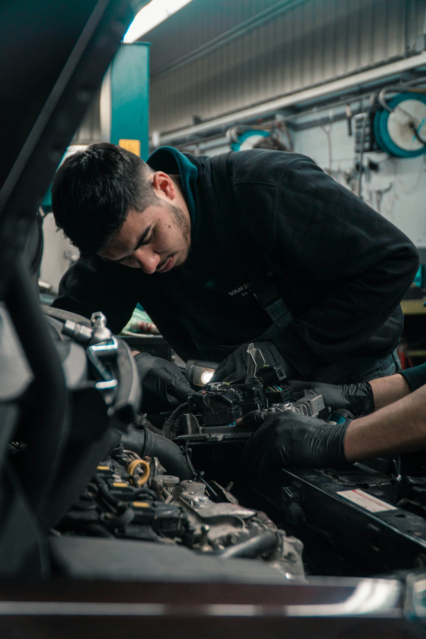 A mechanic in a black hoodie and gloves works on a car engine in a workshop bay