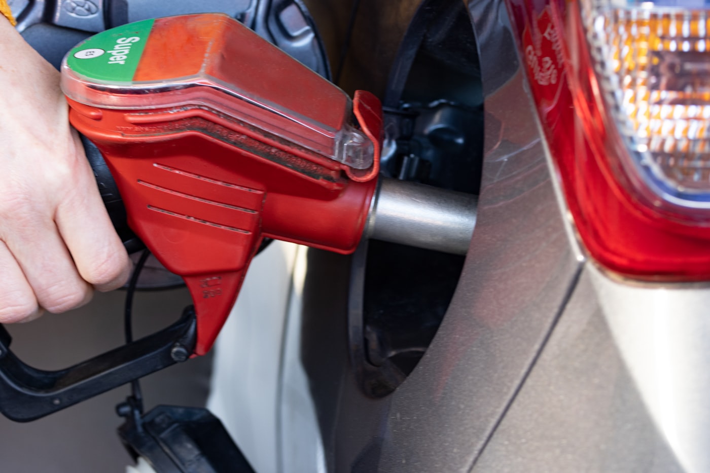 A hand holds a red fuel pump nozzle marked Super, filling a silver car