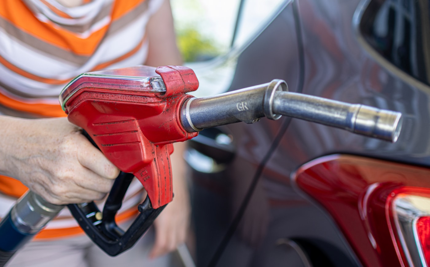 A person in a striped top holds a red fuel nozzle at the filler of a dark grey car