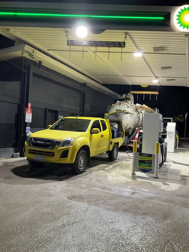 Yellow service ute connected by hose to a large tank at a service station at night, Keith