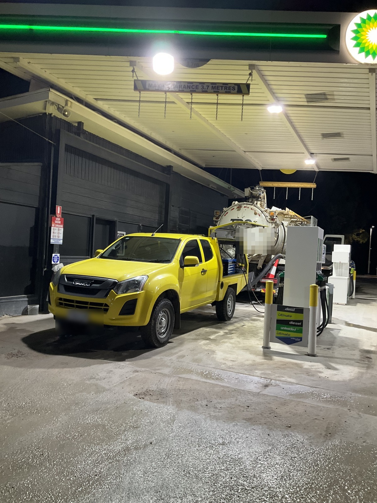 Yellow service ute connected by hose to a large tank at a service station at night, Keith